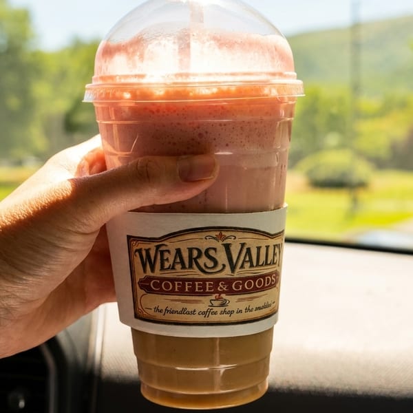 Hand holding a Wears Valley Coffee & Goods drink in a car, with a road and trees outside.