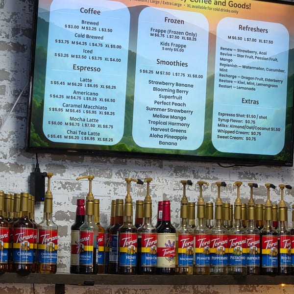 A digital menu board for Wears Valley Coffee and Goods above a shelf of Torani syrup bottles.