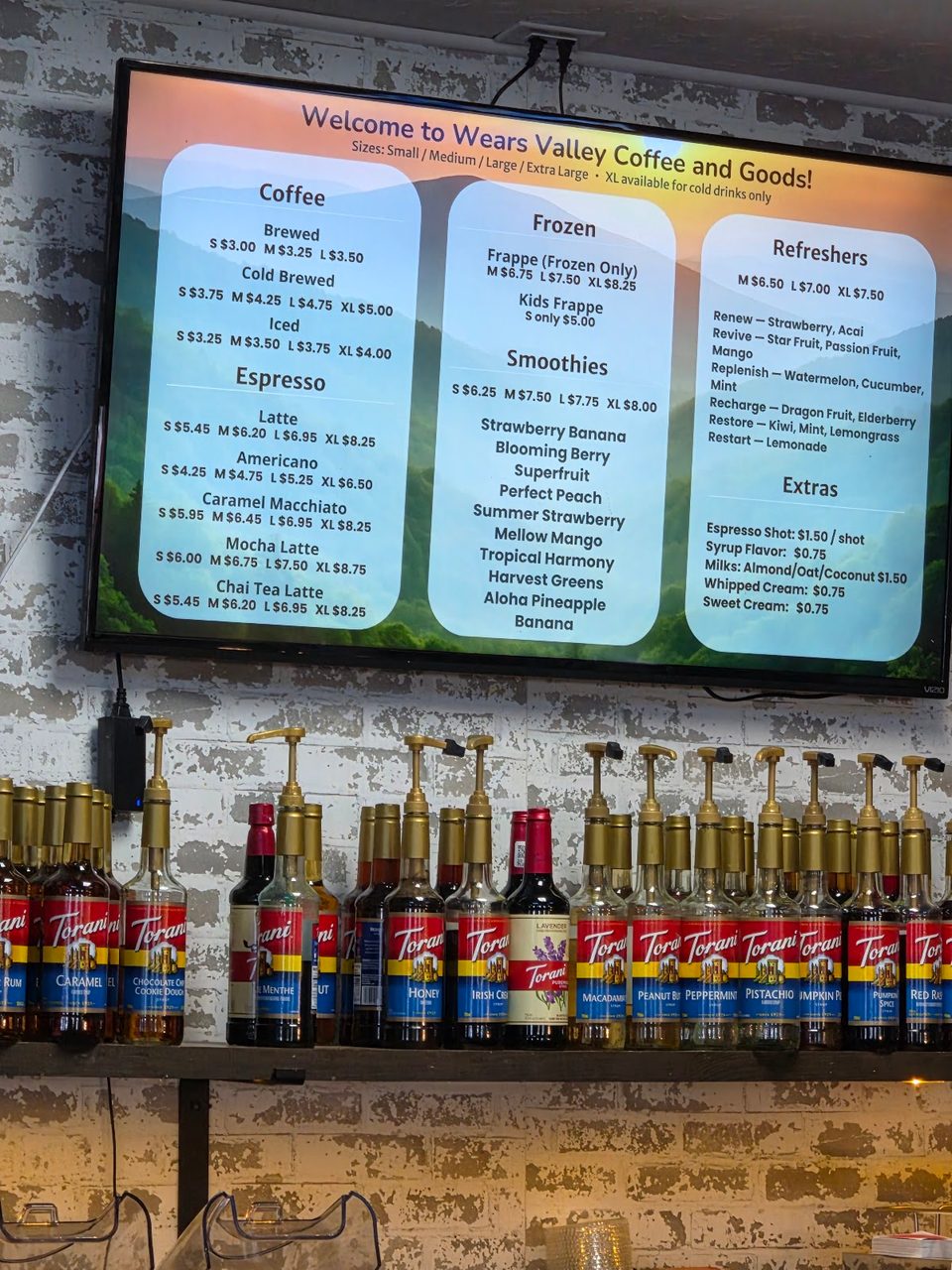 A digital menu board for Wears Valley Coffee and Goods above a shelf of Torani syrup bottles.