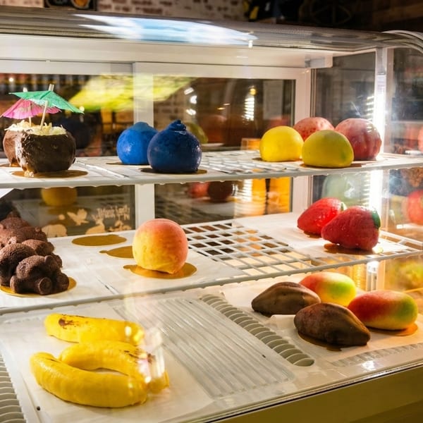 A display case filled with various colorful, fruit-shaped desserts, including chocolate-covered bananas and berries.
